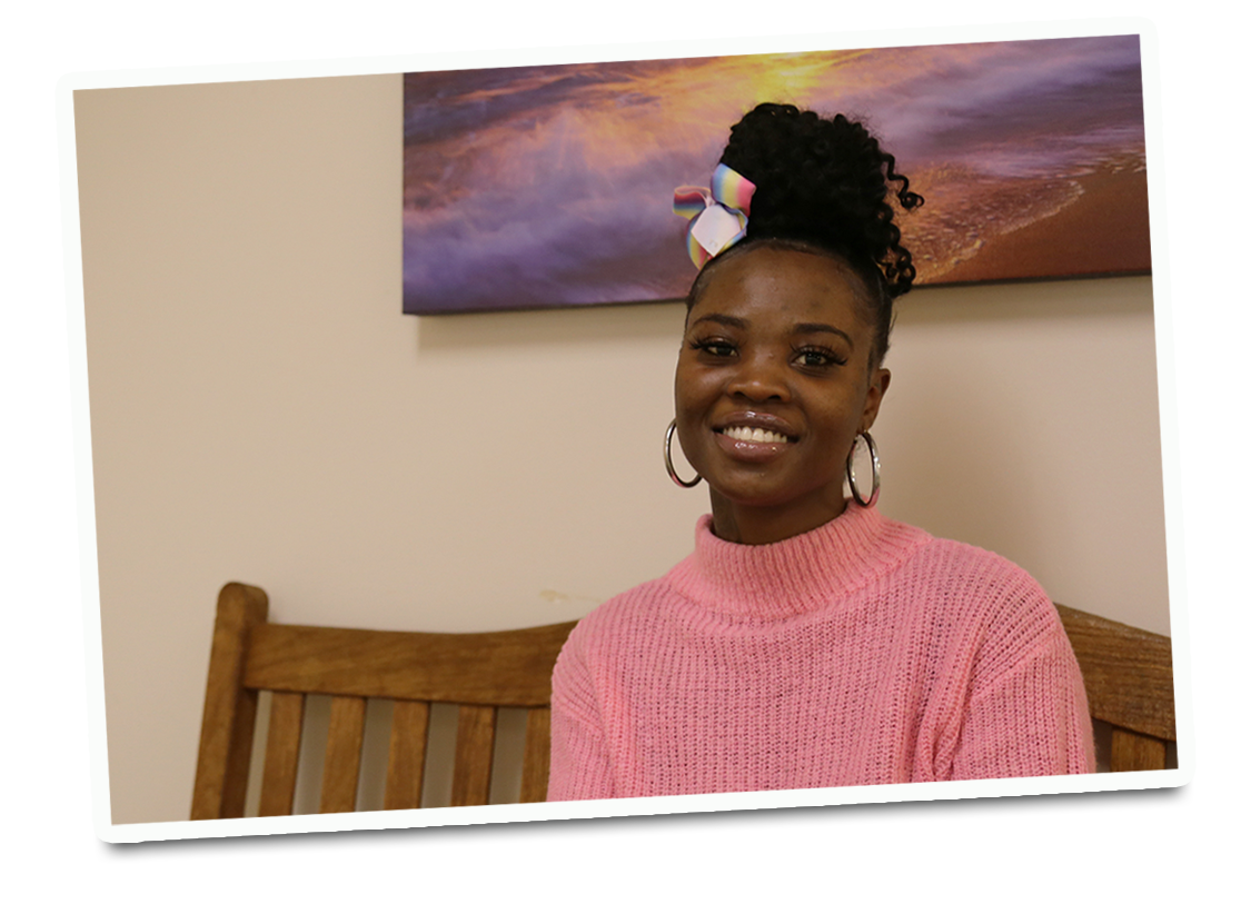 A smiling young, black woman with her hair in a bun on the top of her head wearing a pink turtleneck sweater. 